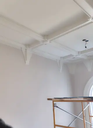 A white coffered ceiling with carved corbels above an arched window, a scaffold still standing below.