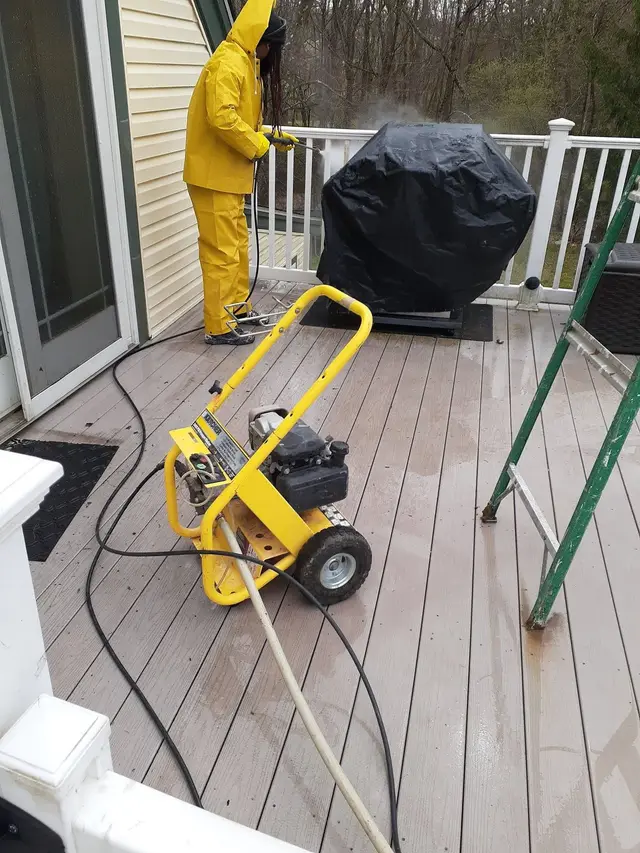 A worker in yellow rain gear pressure-washing a deck beside a covered grill and white railings.
