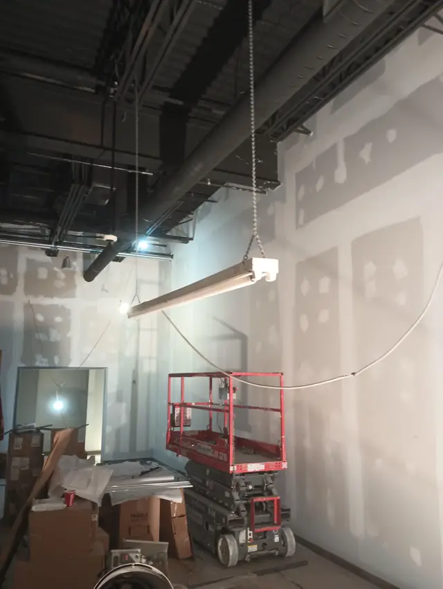 New drywall in a commercial space, its seams taped and screws spotted with compound, a scissor lift beside it.