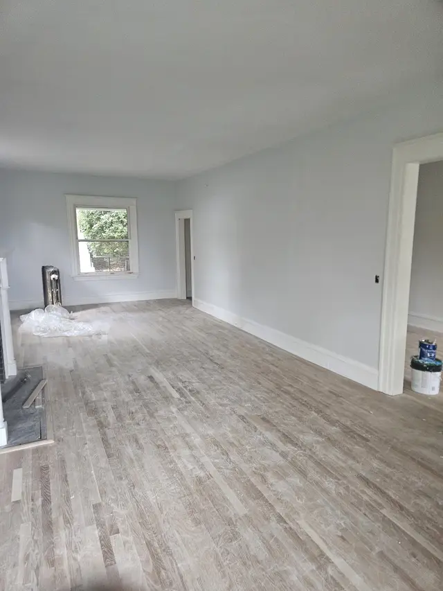 A long empty room with smooth white walls and a pale wood floor, lit by a single window.