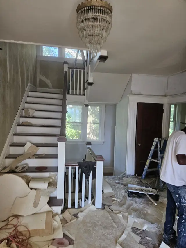 A period foyer and staircase mid-renovation, the floor strewn with stripped material and a painter at work.