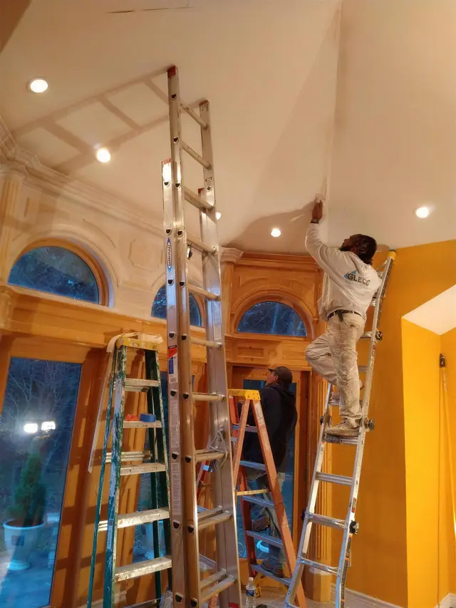 Two painters on extension ladders cutting in a cream ceiling above arched windows and honey wood paneling, beside a marigold wall.