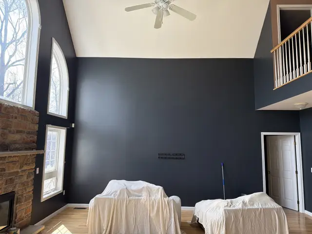 A two-story great room with a deep navy accent wall, tall arched windows, a loft rail and furniture under drop cloths.
