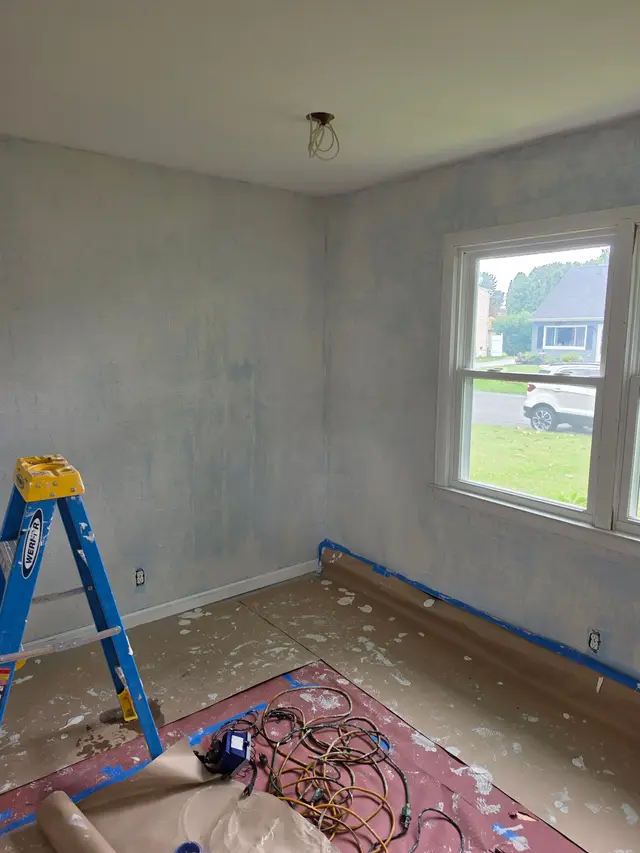 A bedroom with its walls freshly skimmed in gray-white compound, a stepladder and cords on the drop cloth below the window.