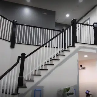 A staircase with black handrails and newel posts, white balusters and black treads, under a tall gray wall.
