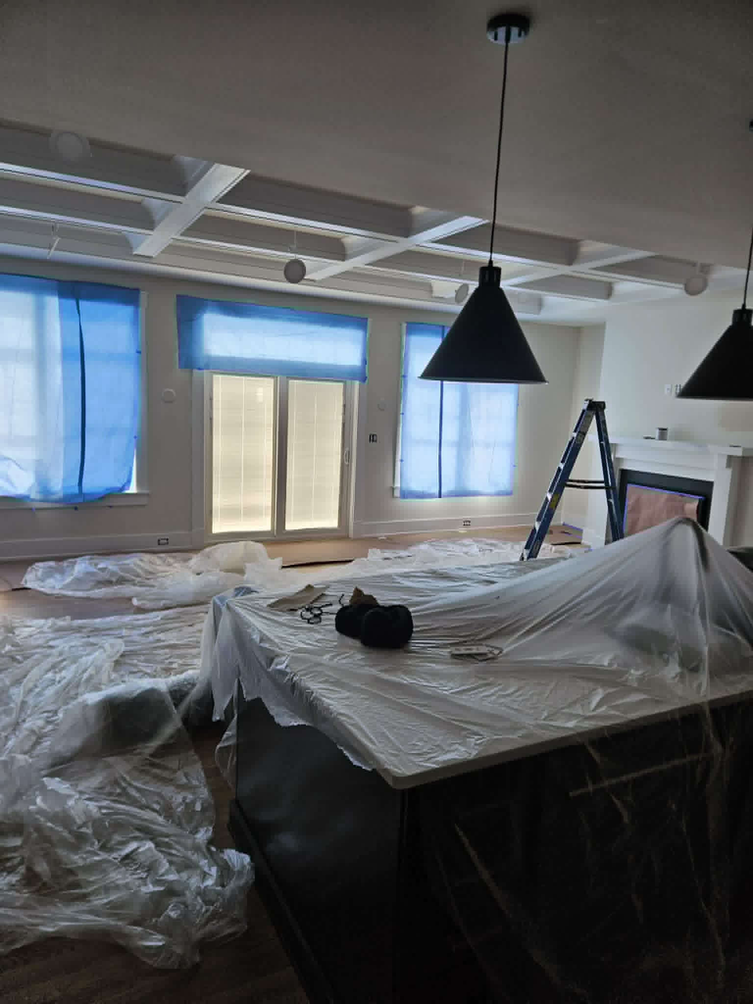 Open living and kitchen space with a white coffered ceiling and black statement pendants, fully draped and masked for painting — interior painting in progress.